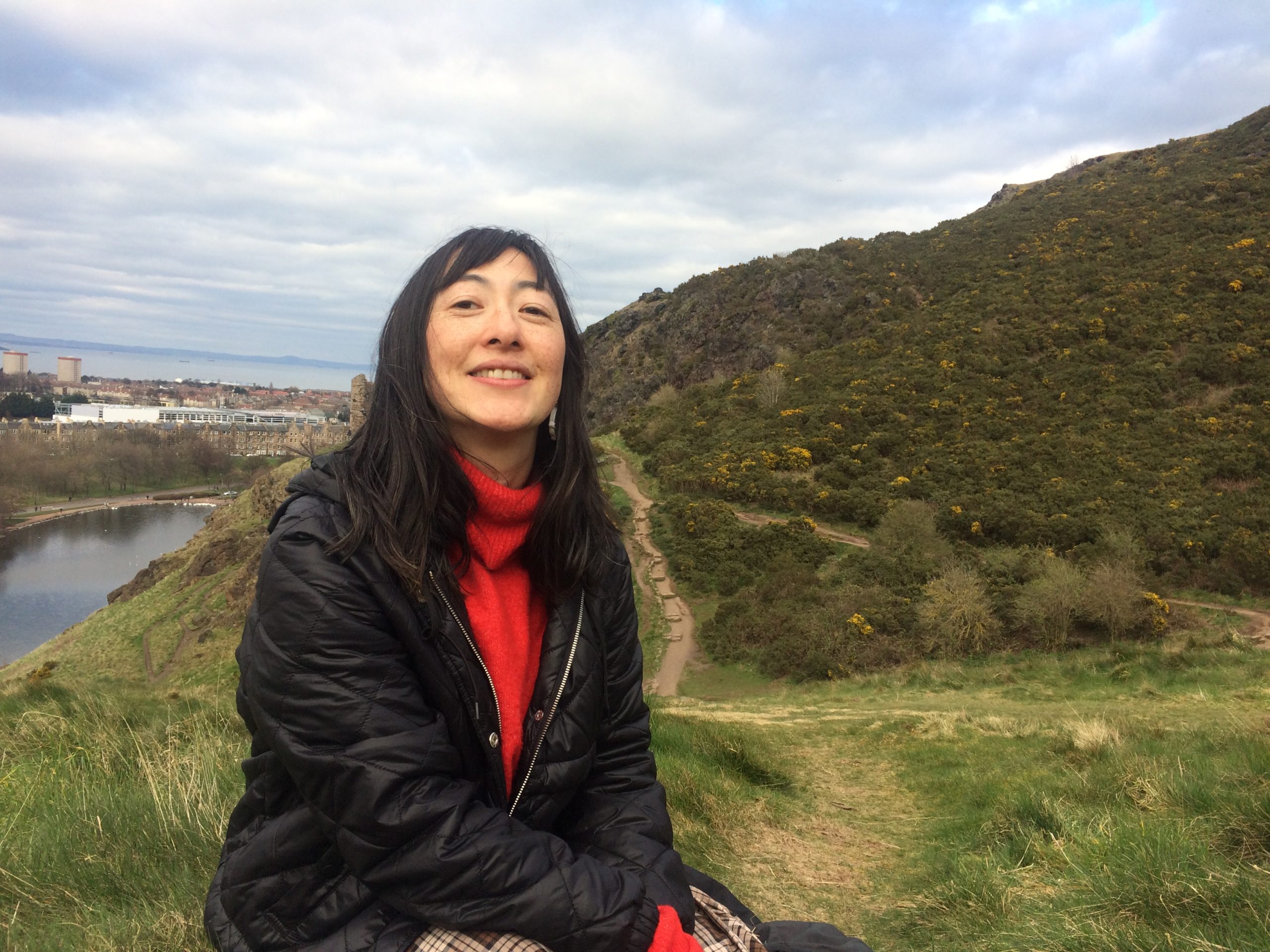 Katch Holmes sits in a red jumper and black coat in front of a green hill, with blue sky and clouds in the background.